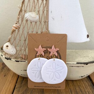 Pode incluir: Brincos de argila branca com um design de d&oacute;lar de areia e um charme de estrela do mar rosa. Os brincos est&atilde;o em um cart&atilde;o marrom com o texto "ENCHANTEDTOKKIROOM.etsy.com".
