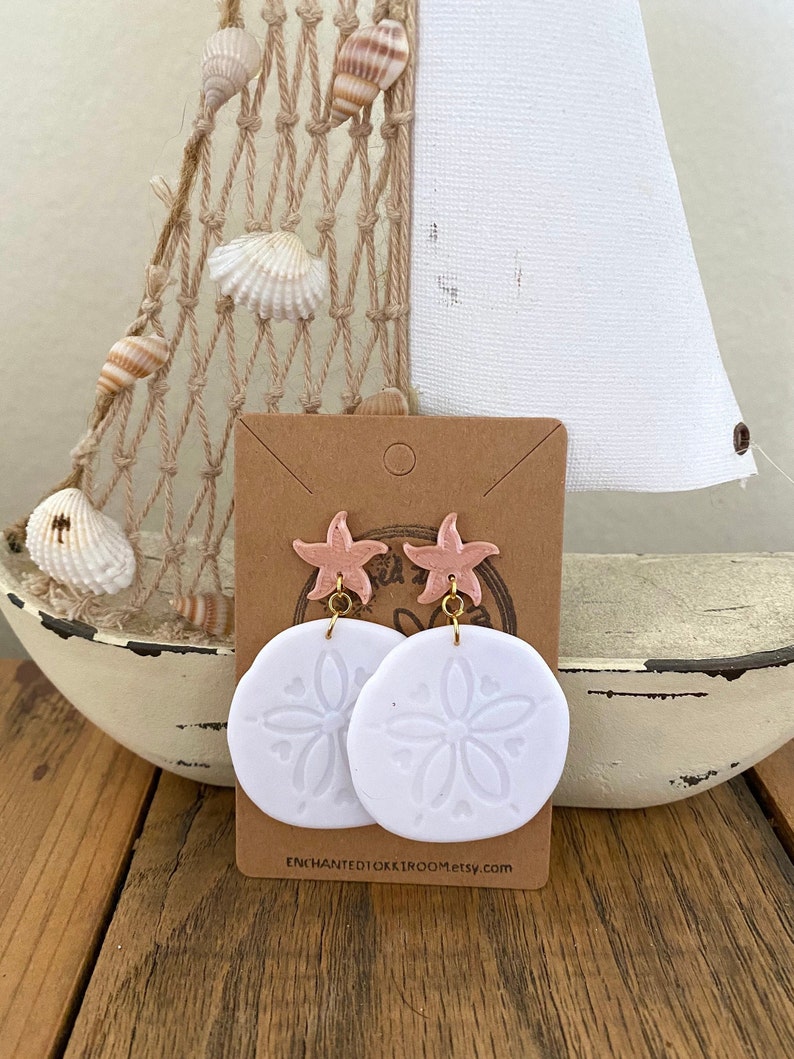 Pode incluir: Brincos de argila branca com um design de d&oacute;lar de areia e um charme de estrela do mar rosa. Os brincos est&atilde;o em um cart&atilde;o marrom com o texto "ENCHANTEDTOKKIROOM.etsy.com".