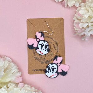 May include: A pair of black and pink cartoon character earrings with gold hooks. The earrings feature a black and white face with a pink bow and a white smile. The earrings are on a brown card with the text "Enchanted Dickens" printed on it.