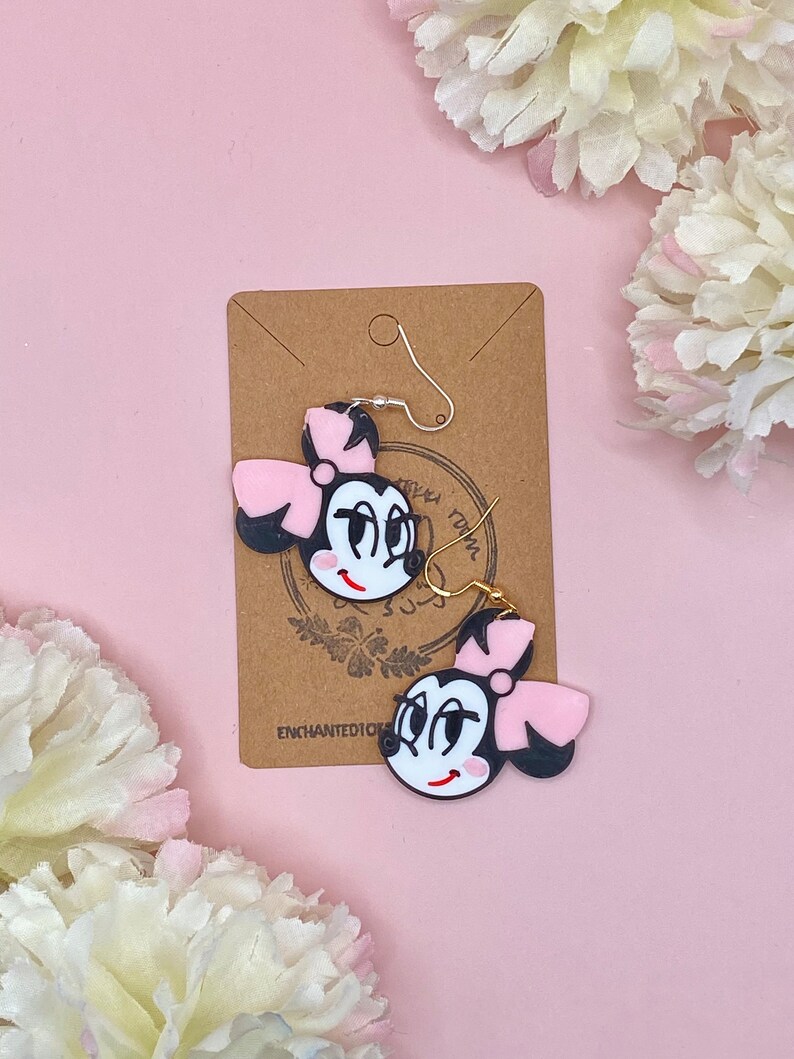 May include: A pair of black and pink cartoon character earrings with gold hooks. The earrings feature a black and white face with a pink bow and a white smile. The earrings are on a brown card with the text "Enchanted Dickens" printed on it.