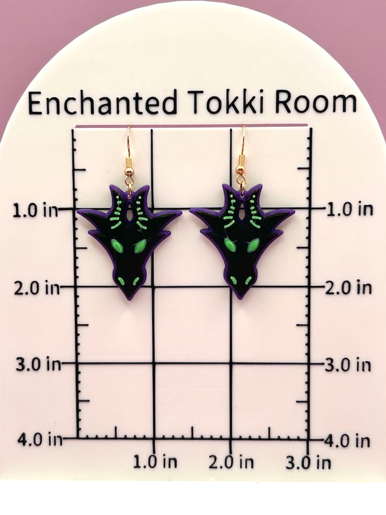 May include: A pair of black and purple earrings with a green dragon design. The earrings are approximately 2 inches long.