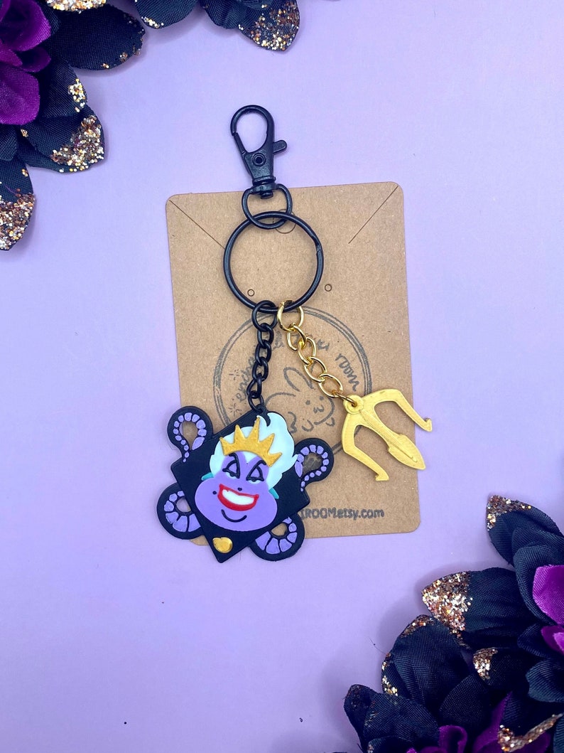 Op de afbeelding: Paarse Ursula sleutelhanger met een gouden drietand bedel en een zwarte ketting. De sleutelhanger is bevestigd aan een zwarte split ring en een zwarte karabijnhaak.