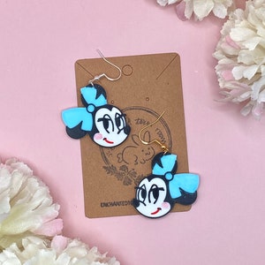 May include: A pair of black and white cartoon character earrings with a blue bow. The earrings are on a brown card with the text "Enchanted101" and a small rabbit logo.