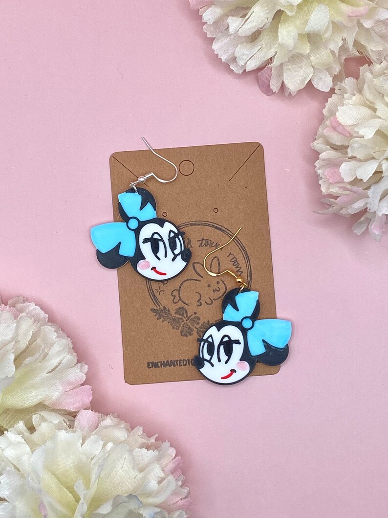 May include: A pair of black and white cartoon character earrings with a blue bow. The earrings are on a brown card with the text "Enchanted101" and a small rabbit logo.