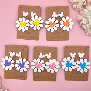 May include: Six pairs of dangle earrings featuring a white daisy with a Mickey Mouse silhouette in the center. The daisies are in various colors, including yellow, orange, purple, pink, blue, and light blue.