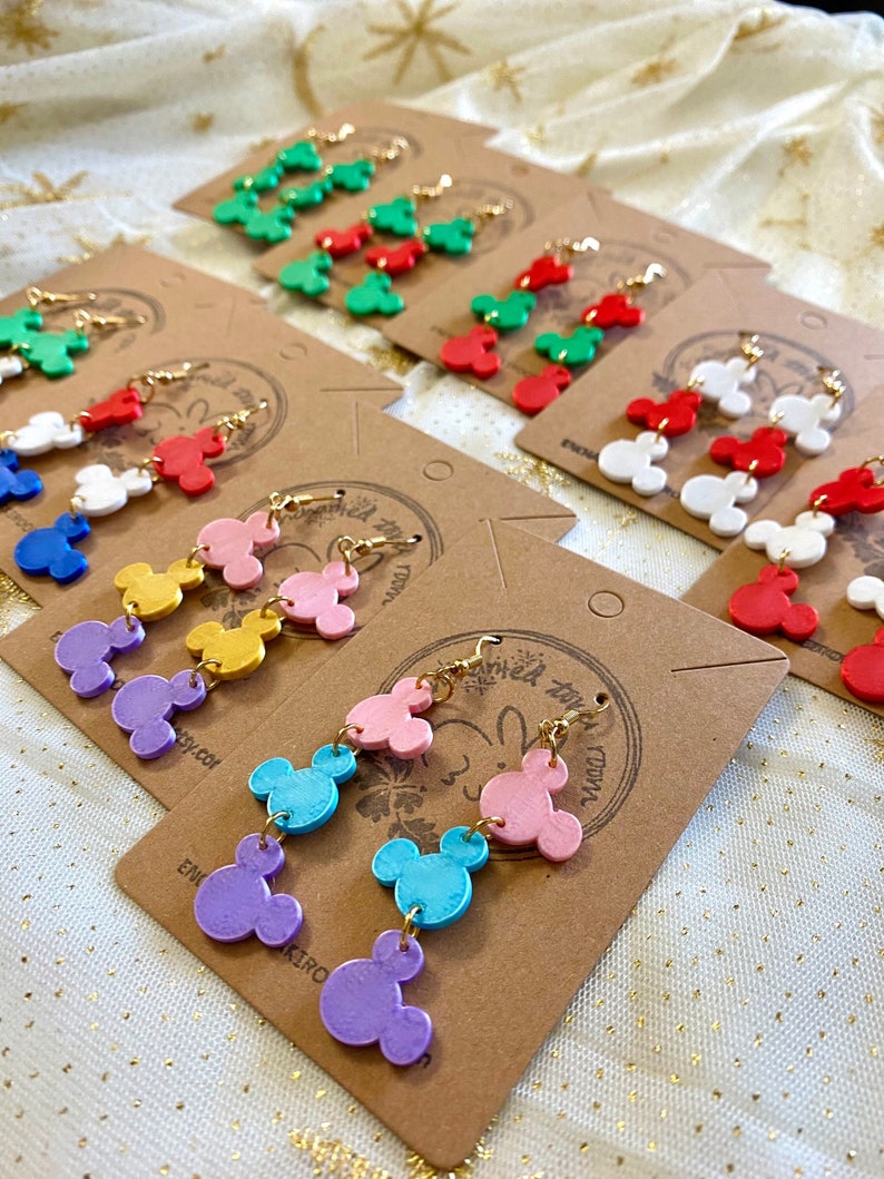 May include: A set of colourful Mickey Mouse-shaped earrings. Each pair features three stacked Mickey Mouse heads in different colours, including purple, blue, pink, yellow, red, green, and white. The earrings are displayed on brown card stock with the text "Enchanted Tiny Town" printed on the card.