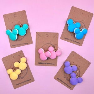 May include: Five pairs of handmade polymer clay earrings shaped like Mickey Mouse ears. The earrings are in various colours, including blue, pink, yellow, and purple. Each pair is displayed on a brown card with the Etsy shop name "enchantedcrookroom.etsy.com".