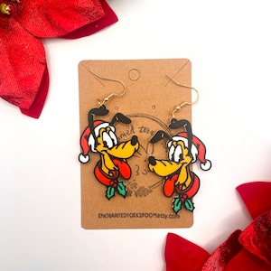 Puede incluir: Un par de pendientes colgantes festivos con un personaje de perro de dibujos animados que lleva un gorro de Papá Noel y un collar rojo con acentos de acebo. Los pendientes están sujetos a ganchos dorados y se muestran en una tarjeta marrón. El texto "ENCHANTEDIOKIRROOMetsy.com" es visible.