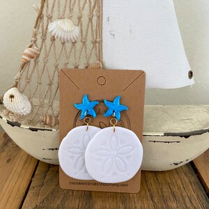 Pode incluir: Brincos de d&oacute;lar de areia branco com um charme de estrela do mar azul. Os brincos est&atilde;o em um cart&atilde;o marrom com o texto "ENCHANTED10KROOM.etsy.com".
