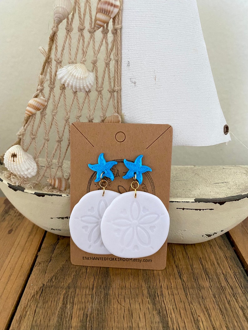 Pode incluir: Brincos de d&oacute;lar de areia branco com um charme de estrela do mar azul. Os brincos est&atilde;o em um cart&atilde;o marrom com o texto "ENCHANTED10KROOM.etsy.com".
