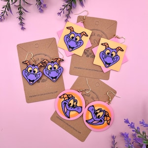 Può includere: Tre paia di orecchini pendenti con un drago viola di cartone animato con corna arancioni. Gli orecchini sono su uno sfondo marrone con il testo "enchantedicksbroom.etsy.com".