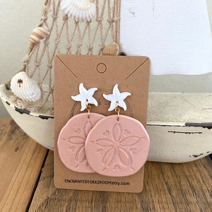 Pode incluir: Um par de brincos pendentes rosa e branco com um charme de estrela do mar e um design de d&oacute;lar de areia. Os brincos s&atilde;o exibidos em um cart&atilde;o marrom com o texto "ENCHANTEDTOKKIROOMetsy.com".