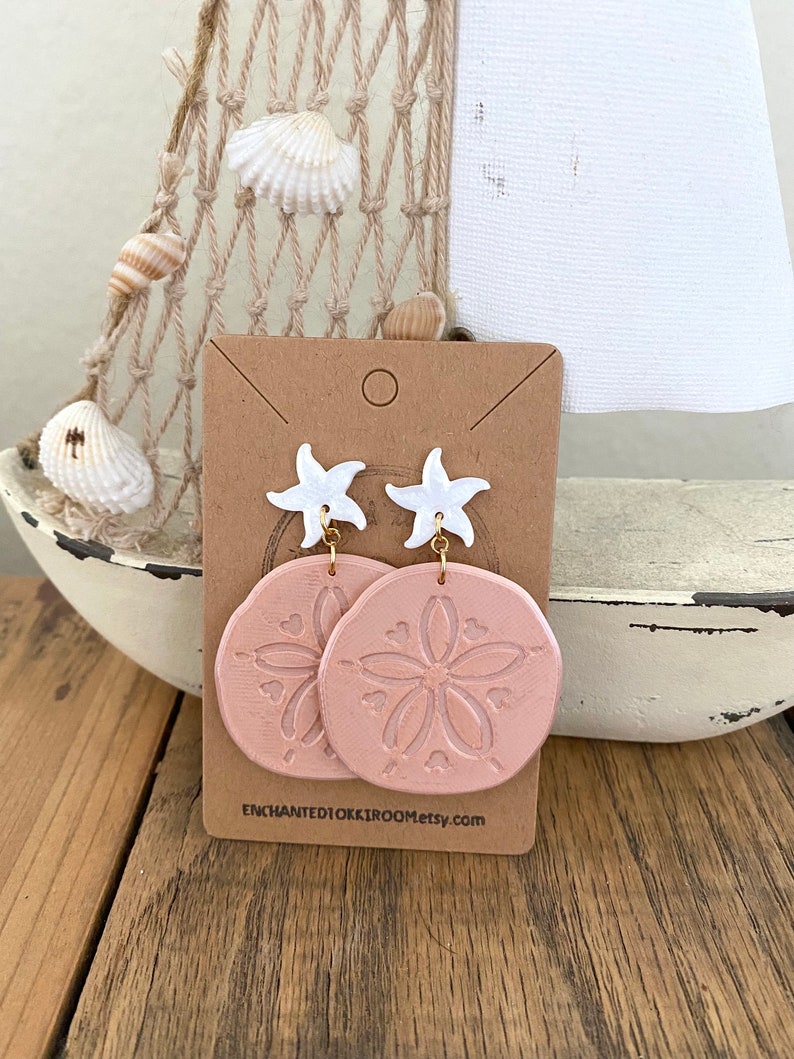 Pode incluir: Um par de brincos pendentes rosa e branco com um charme de estrela do mar e um design de d&oacute;lar de areia. Os brincos s&atilde;o exibidos em um cart&atilde;o marrom com o texto "ENCHANTEDTOKKIROOMetsy.com".