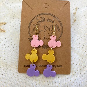 May include: A pair of dangle earrings with three layered charms. The charms are shaped like Mickey Mouse heads and are painted in pink, gold, and purple.