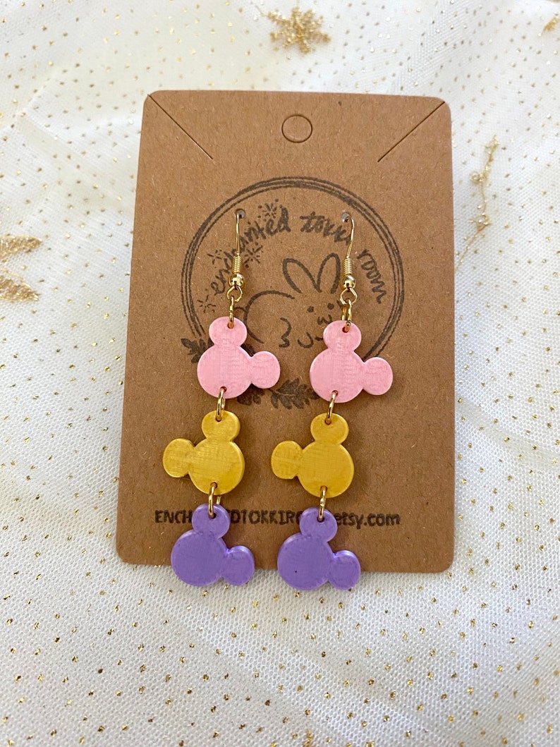 May include: A pair of dangle earrings with three layered charms. The charms are shaped like Mickey Mouse heads and are painted in pink, gold, and purple.