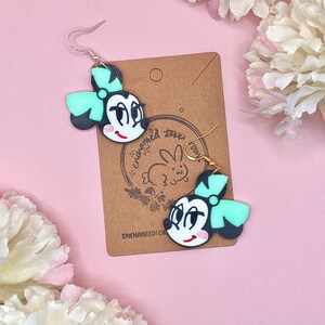 May include: A pair of black and white cartoon character earrings with a green bow. The earrings are on a brown card with the text "enchanted toys" and a rabbit logo.