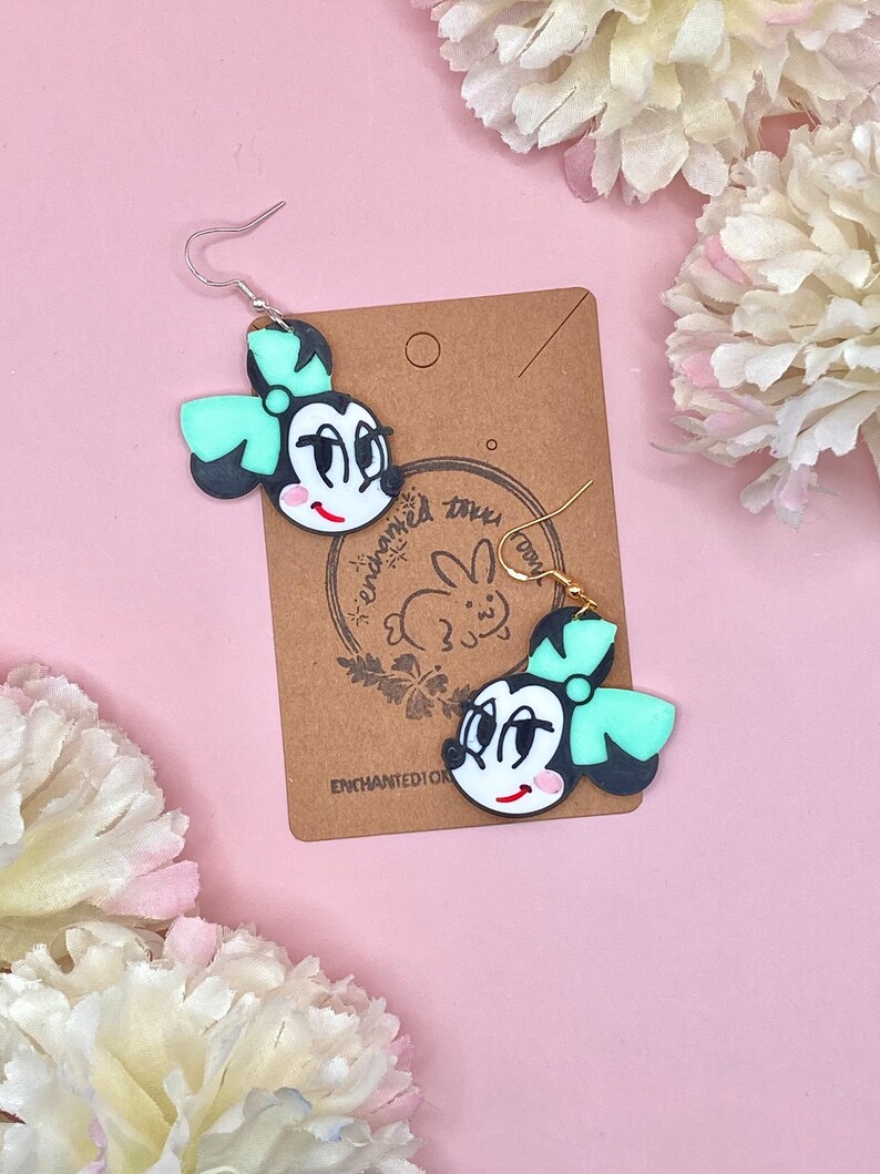 May include: A pair of black and white cartoon character earrings with a green bow. The earrings are on a brown card with the text "enchanted toys" and a rabbit logo.
