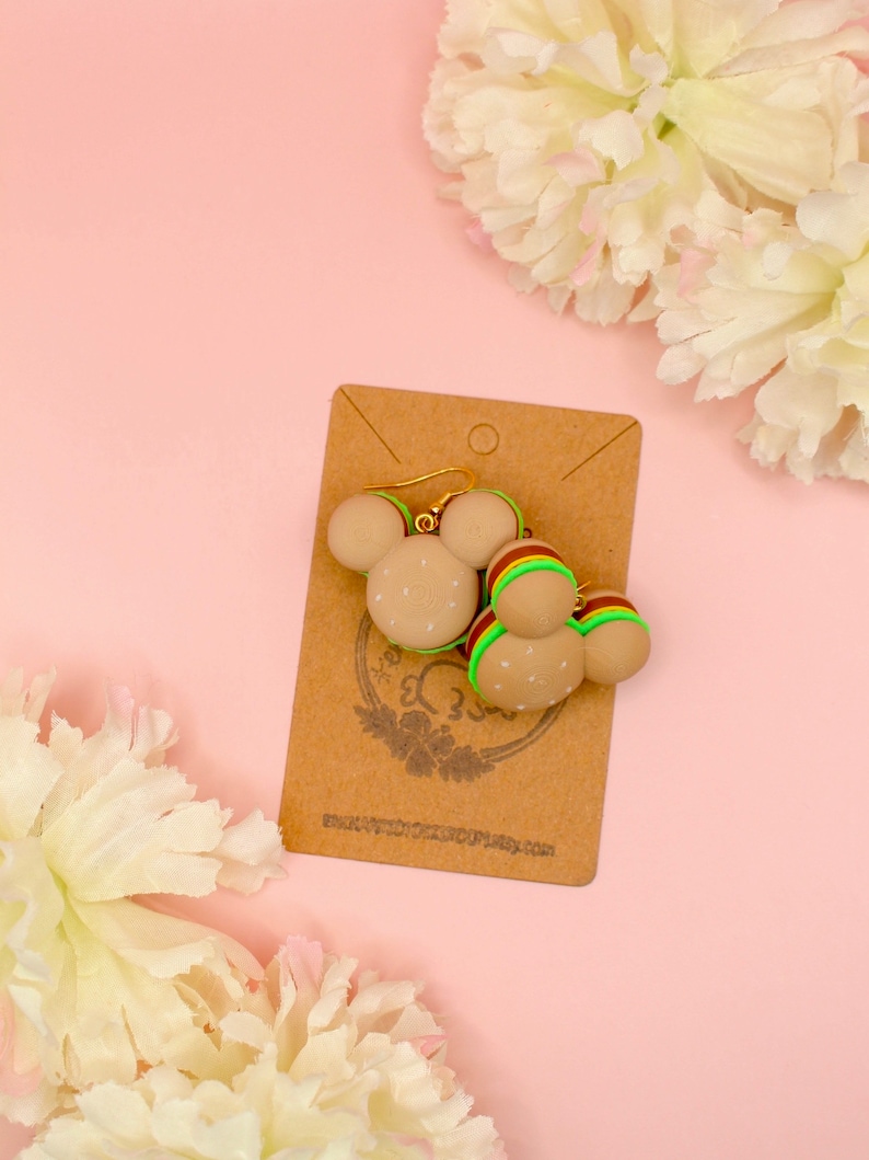 May include: A pair of gold dangle earrings with a brown and green hamburger charm in the shape of Mickey Mouse's head. The earrings are displayed on a brown card with the text "Etsy Shop" and a website address.