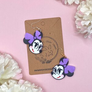 May include: Purple and black cartoon character earrings with a bow. The earrings are on a brown card with the text "Enchanted10k1r001" and a small bunny illustration.