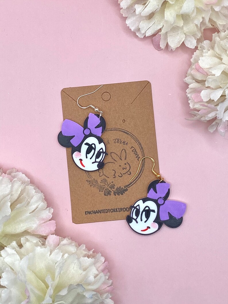 May include: Purple and black cartoon character earrings with a bow. The earrings are on a brown card with the text "Enchanted10k1r001" and a small bunny illustration.