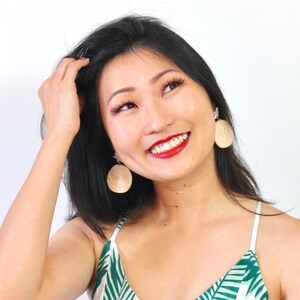 Pode incluir: Uma mulher com cabelo preto comprido est&aacute; sorrindo e usando um top sem mangas de estampa tropical branca e verde com grandes brincos redondos bege.