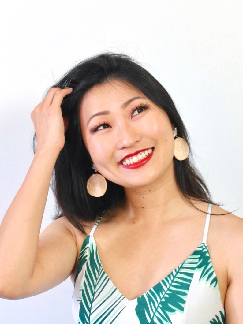 Pode incluir: Uma mulher com cabelo preto comprido est&aacute; sorrindo e usando um top sem mangas de estampa tropical branca e verde com grandes brincos redondos bege.