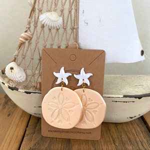 Pode incluir: Um par de brincos de d&oacute;lar de areia cor de p&ecirc;ssego com pingentes de estrela do mar brancos e ganchos dourados. Os brincos s&atilde;o exibidos em um cart&atilde;o marrom com o texto "enchantedtoktroom.etsy.com".