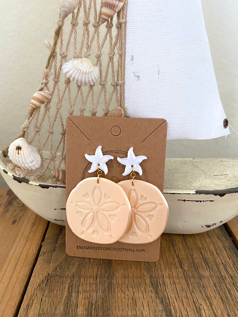 Pode incluir: Um par de brincos de d&oacute;lar de areia cor de p&ecirc;ssego com pingentes de estrela do mar brancos e ganchos dourados. Os brincos s&atilde;o exibidos em um cart&atilde;o marrom com o texto "enchantedtoktroom.etsy.com".