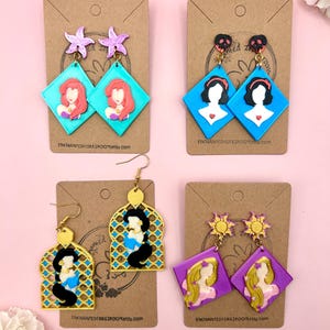 Puede incluir: Cuatro pares de pendientes con temática de princesas. El primer par presenta a Ariel de La Sirenita con un amuleto de estrella de mar púrpura. El segundo par presenta a Blancanieves con un amuleto de corazón rojo. El tercer par presenta a Jasmine de Aladdin con un diseño de filigrana dorada. El cuarto par presenta a Rapunzel con un amuleto de sol dorado.