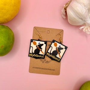 May include: A pair of black and white earrings with a rat holding a wooden spoon and the word "Ratatouille" on each earring. The earrings are on a brown card with the text "EnchantingCrochetery.com".