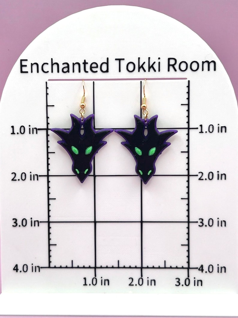 May include: A pair of purple and black dragon earrings with green eyes. The earrings are approximately 1.5 inches long and are hanging from gold hooks.