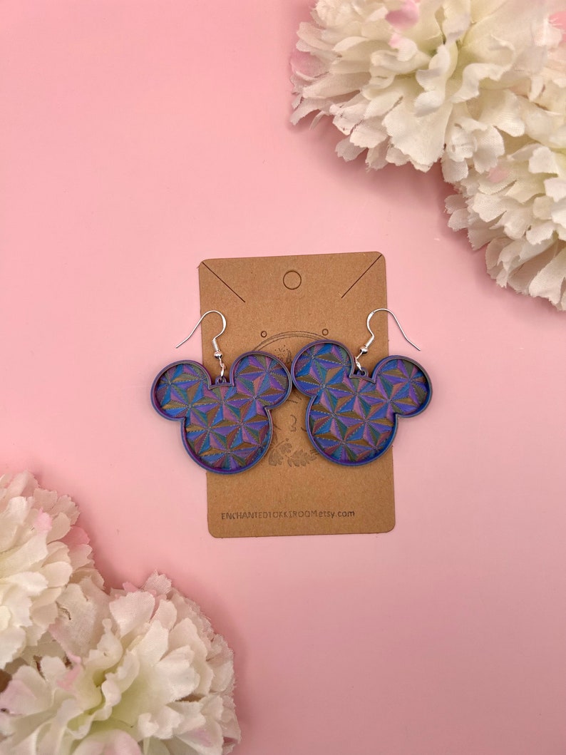 K&ouml;nnte beinhalten: Ohrringe in Mickey Mouse-Form mit einem geometrischen Muster in Blau, Lila und Gr&uuml;n. Die Ohrringe h&auml;ngen an silbernen Haken und werden auf einer braunen Karte pr&auml;sentiert. Der Hintergrund ist hellrosa.