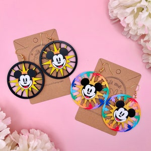 May include: A pair of colorful Mickey Mouse earrings. The earrings are made of wood and feature a Mickey Mouse face in the center of a sunburst design. One pair is black and yellow, the other is rainbow colored.