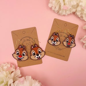 Puede incluir: Un par de pendientes de chipmunk de dibujos animados marrones y blancos en una tarjeta marrón con el texto "enchanteddicefrometsy.com".