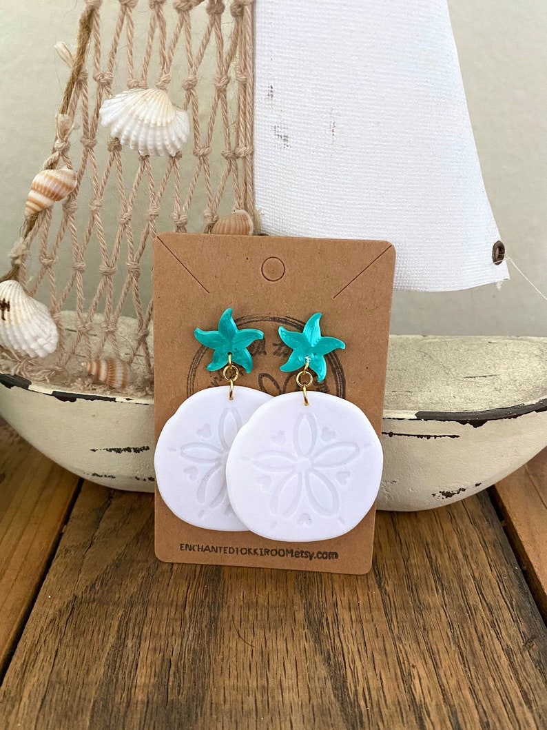 Pode incluir: Brincos de argila branca com um charme de estrela do mar turquesa e um design de d&oacute;lar de areia. Os brincos est&atilde;o em um cart&atilde;o marrom com o texto "ENCHANTED10KK1ROOMetsy.com".
