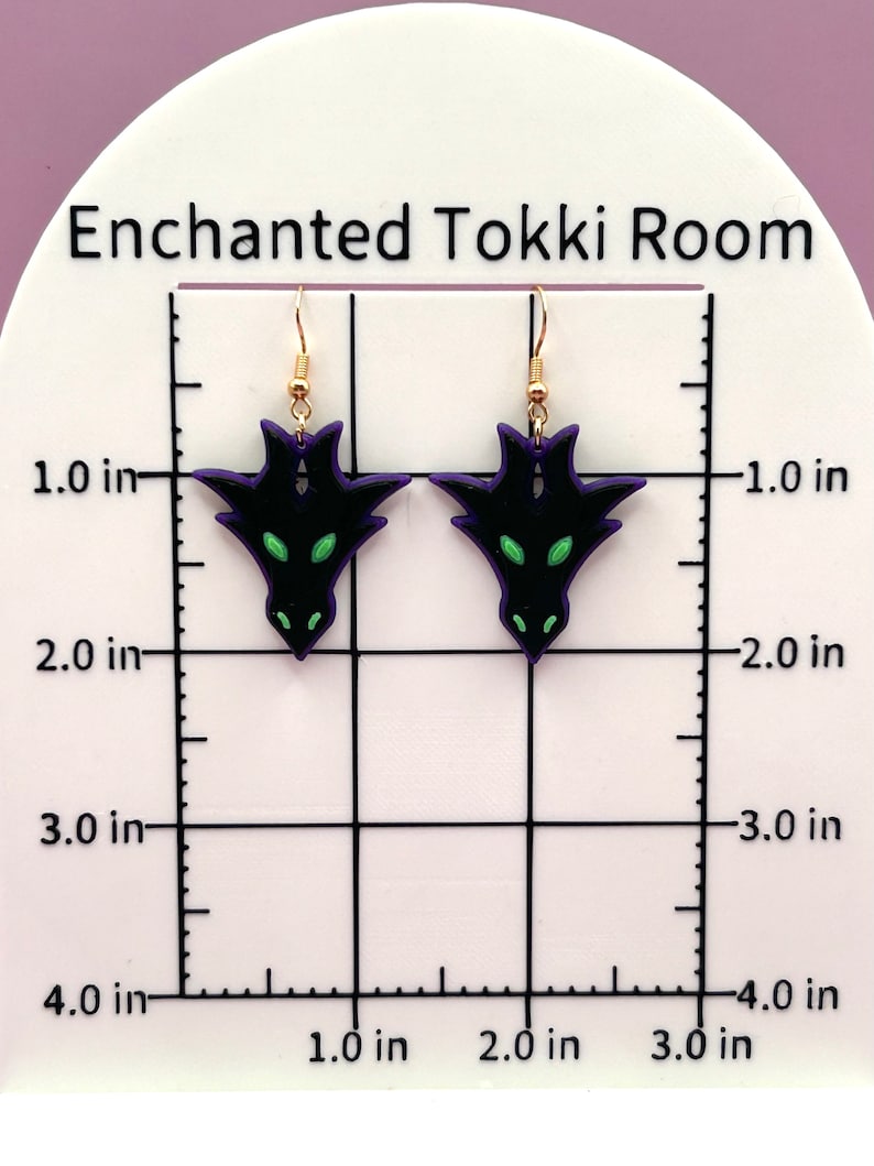 May include: A pair of black and purple dragon earrings with green eyes. The earrings are approximately 1.5 inches long and are hanging from gold hooks.