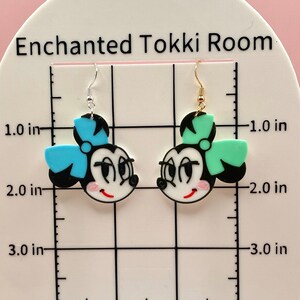 May include: A pair of dangle earrings featuring cartoon faces with black hair, white faces, red lips, and large eyes. One earring has a blue bow and the other has a green bow. The earrings are approximately 5 cm long.