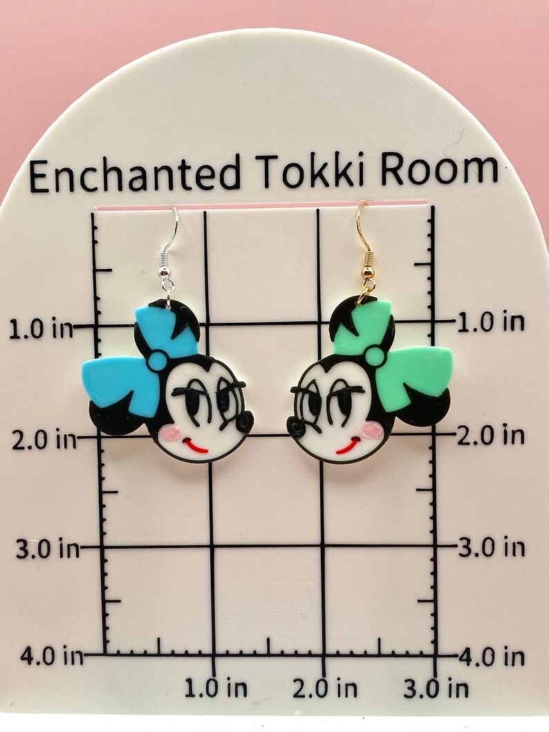 May include: A pair of dangle earrings featuring cartoon faces with black hair, white faces, red lips, and large eyes. One earring has a blue bow and the other has a green bow. The earrings are approximately 5 cm long.