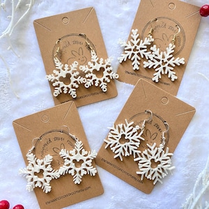 May include: Three pairs of white snowflake earrings with gold hoops. The earrings are displayed on brown card stock with the text "Enchanted Toki Room" and a website address.