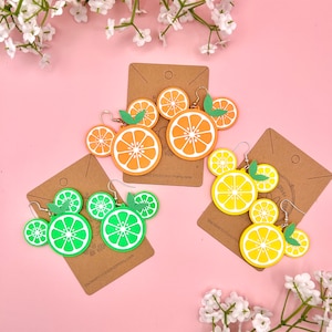 May include: Three pairs of fruit-shaped earrings. The first pair is orange slices, the second pair is lime slices, and the third pair is lemon slices. The earrings are made of wood and painted in bright colors.