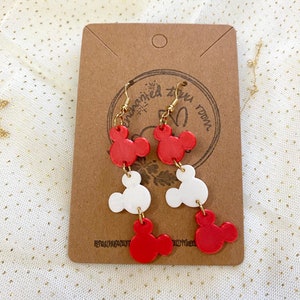 May include: Red and white Mickey Mouse head dangle earrings with gold hooks. The earrings are displayed on a brown card with the text "Dream Big" in white.