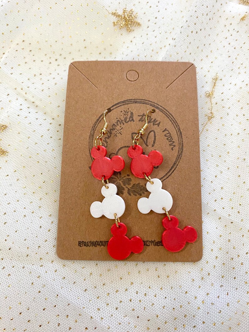 May include: Red and white Mickey Mouse head dangle earrings with gold hooks. The earrings are displayed on a brown card with the text "Dream Big" in white.