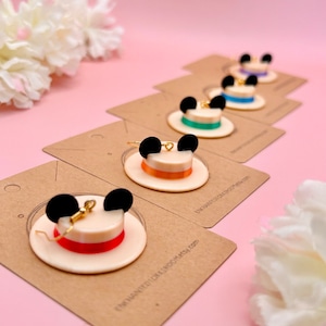 May include: A set of six pairs of earrings featuring a white hat with black Mickey Mouse ears and a colorful ribbon. The earrings are displayed on brown cards with the text "Enchanted Charm Shop Etsy.com".