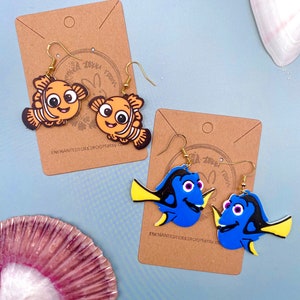 Puede incluir: Un par de pendientes colgantes con personajes de dibujos animados de la película Buscando a Nemo. El primer par presenta un pez payaso amarillo y naranja con rayas negras. El segundo par presenta un pez cirujano azul con aletas amarillas y marcas negras. Ambos pares tienen ganchos dorados.