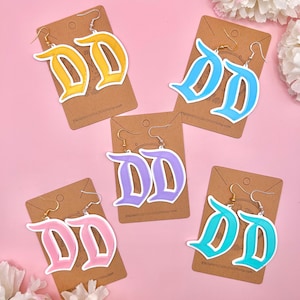 May include: Four pairs of dangle earrings featuring the letter "D" in a gothic font. The earrings are made of white acrylic and are available in yellow, light blue, lavender, and turquoise.