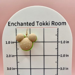 May include: A pair of green, white, and brown burger-shaped earrings with gold hooks. The earrings are about 1 inch in diameter and are shown on a white grid with inch markings.