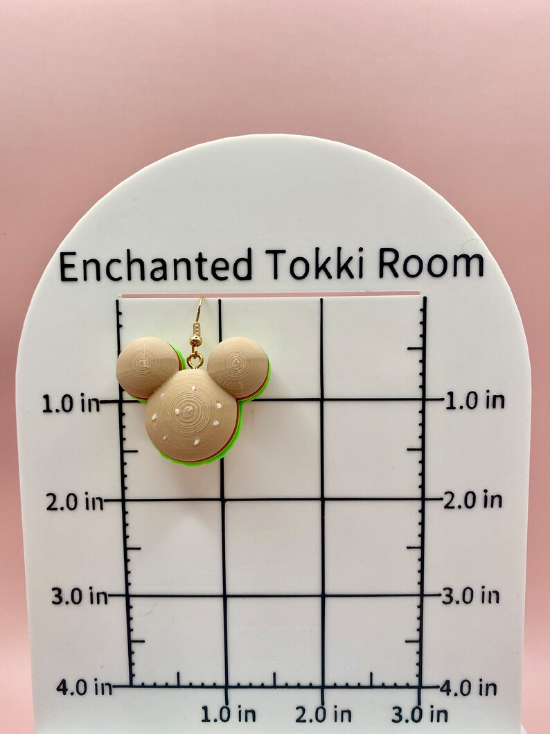 May include: A pair of green, white, and brown burger-shaped earrings with gold hooks. The earrings are about 1 inch in diameter and are shown on a white grid with inch markings.