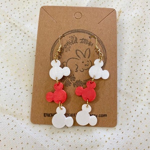 May include: A pair of dangle earrings featuring three white and two red Mickey Mouse head charms. The charms are attached to gold-coloured hoops.