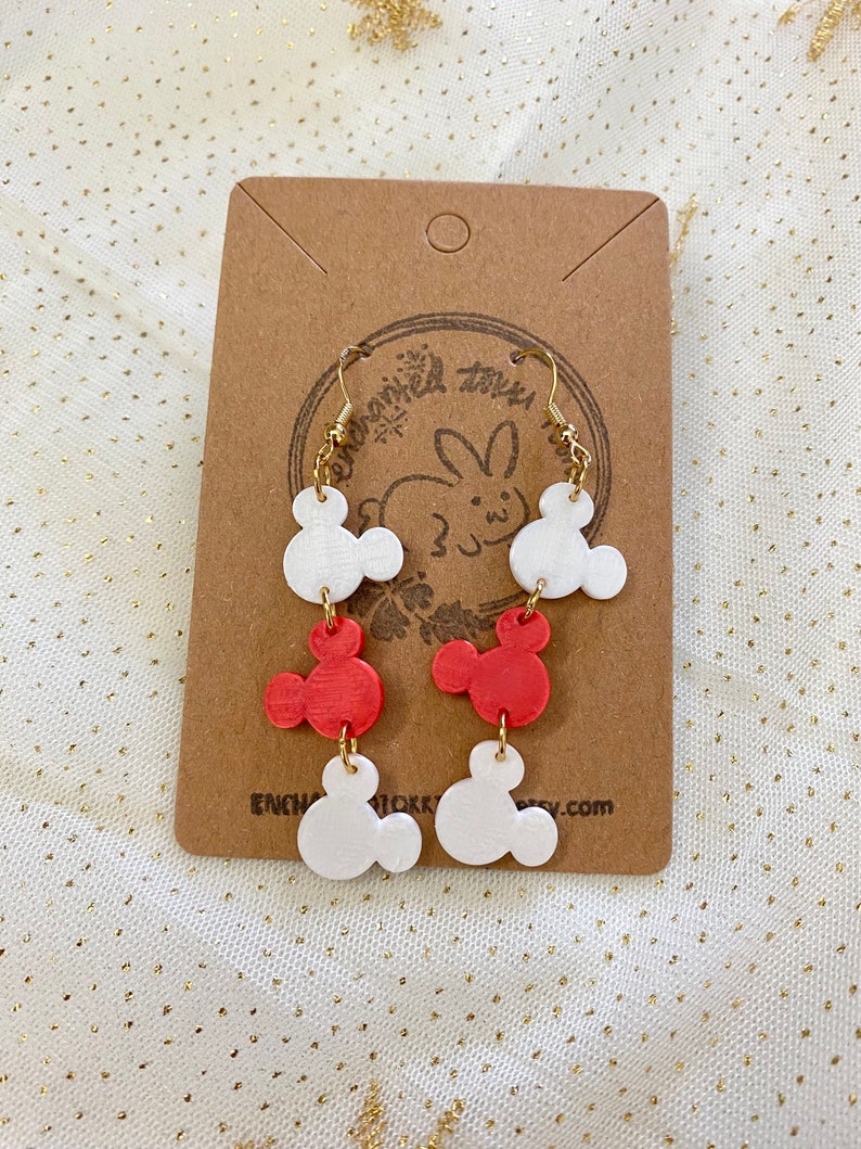May include: A pair of dangle earrings featuring three white and two red Mickey Mouse head charms. The charms are attached to gold-coloured hoops.