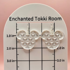 K&ouml;nnte beinhalten: Klare Ohrringe in Form eines Mickey Mouse-Kopfes aus Acryl mit einem geometrischen Muster. Die Ohrringe werden auf einem wei&szlig;en St&auml;nder mit dem Text "Enchanted Tokki Room" pr&auml;sentiert. Die Ohrringe sind etwa 5 cm hoch.